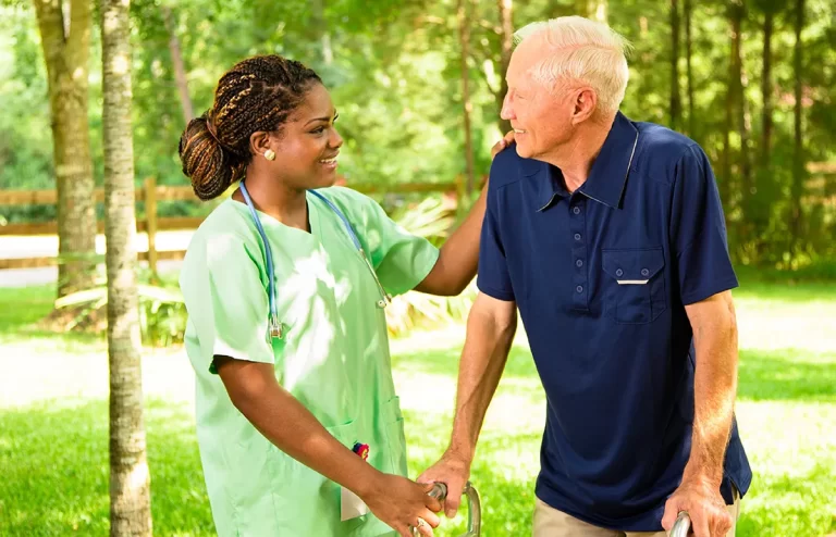 A personal support worker in green scrubs offering support to an elderly man using a walker outdoors